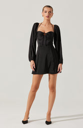 A woman is wearing a black dress with a fitted, strapless bodice featuring a keyhole cutout at the center and thin shoulder straps that tie at the front. The dress has long, slightly puffed sleeves made of a shiny, satin-like fabric.